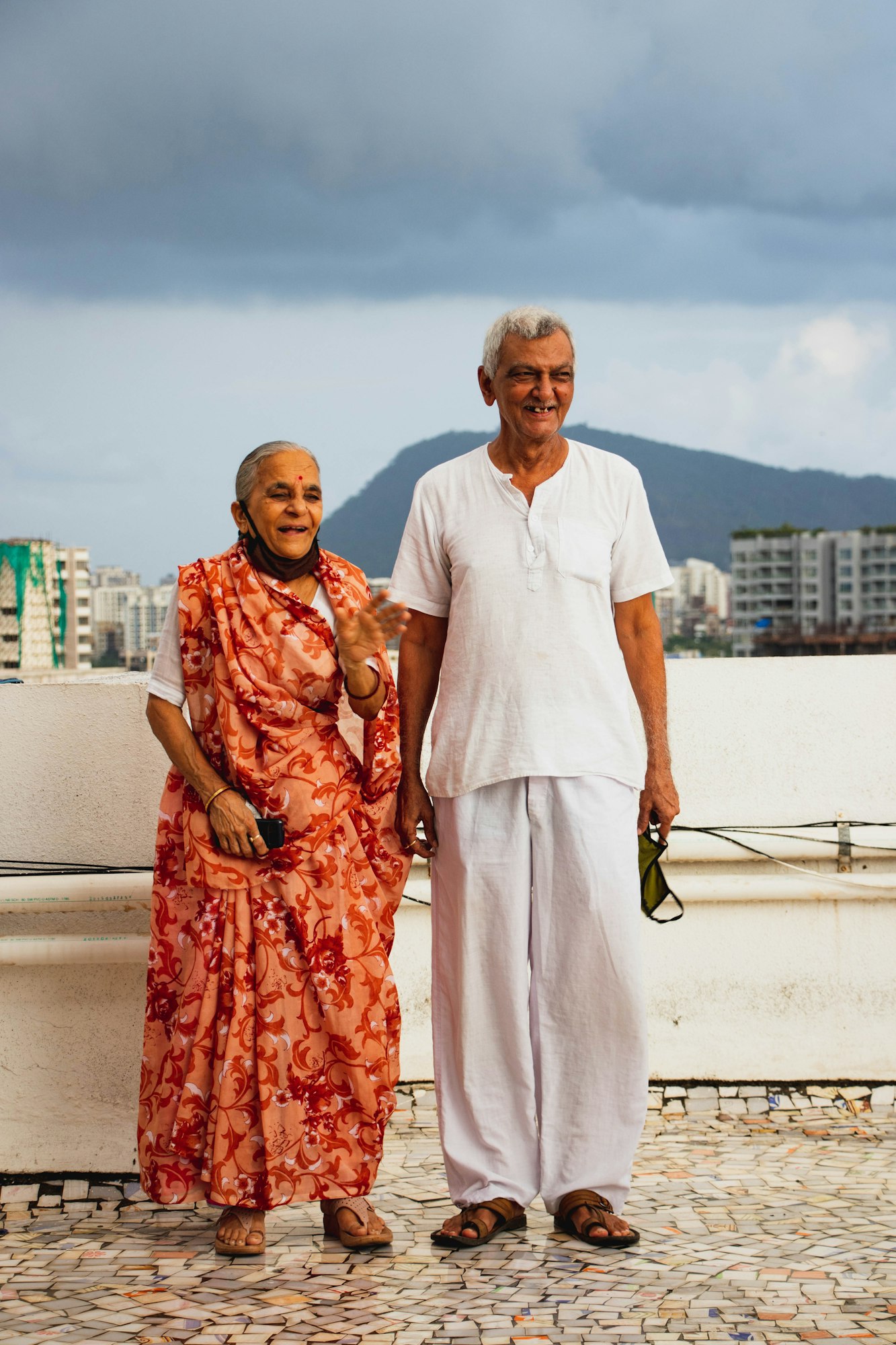 An elderly couple, she in an orange sari and he in a white kurta, smiling together on a rooftop